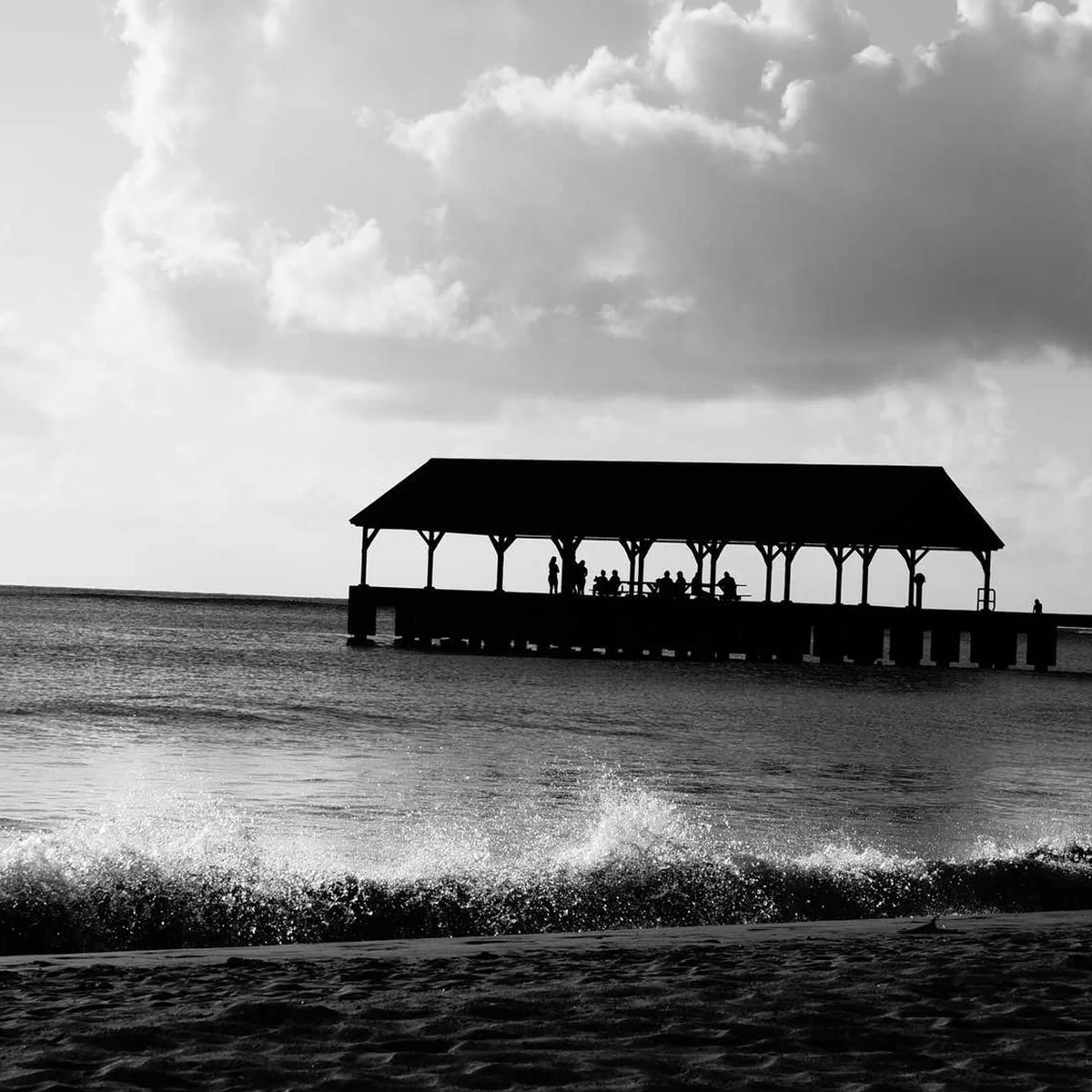 Black and white silhouette of beach pavilion pier with people against ocean and cloudy sky