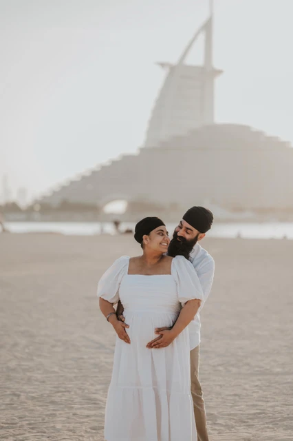 An expecting couple stands together on a Dubai beach with Burj Al Arab visible in the background.