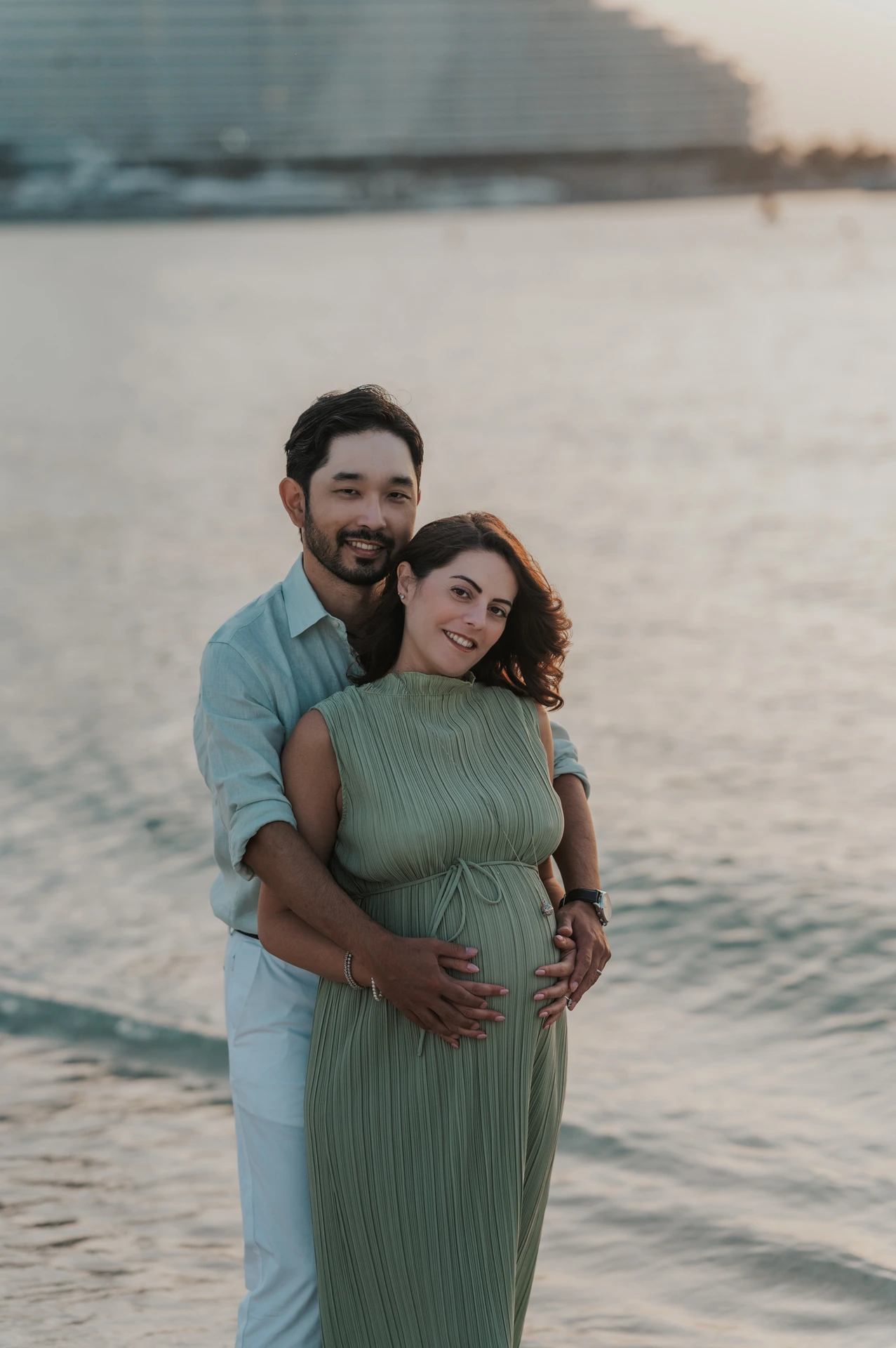 Pregnant woman and partner embrace at Dubai beach shoreline during sunset, capturing a relaxed maternity babymoon couple portrait session.