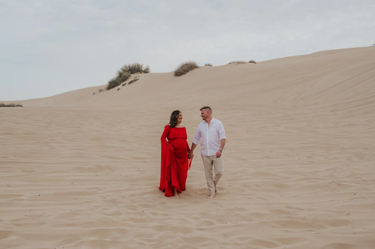 Babymoon photoshoot Dubai dunes Pregnant woman in a flowing red dress walks with partner across Dubai desert dunes during a relaxed maternity photoshoot.