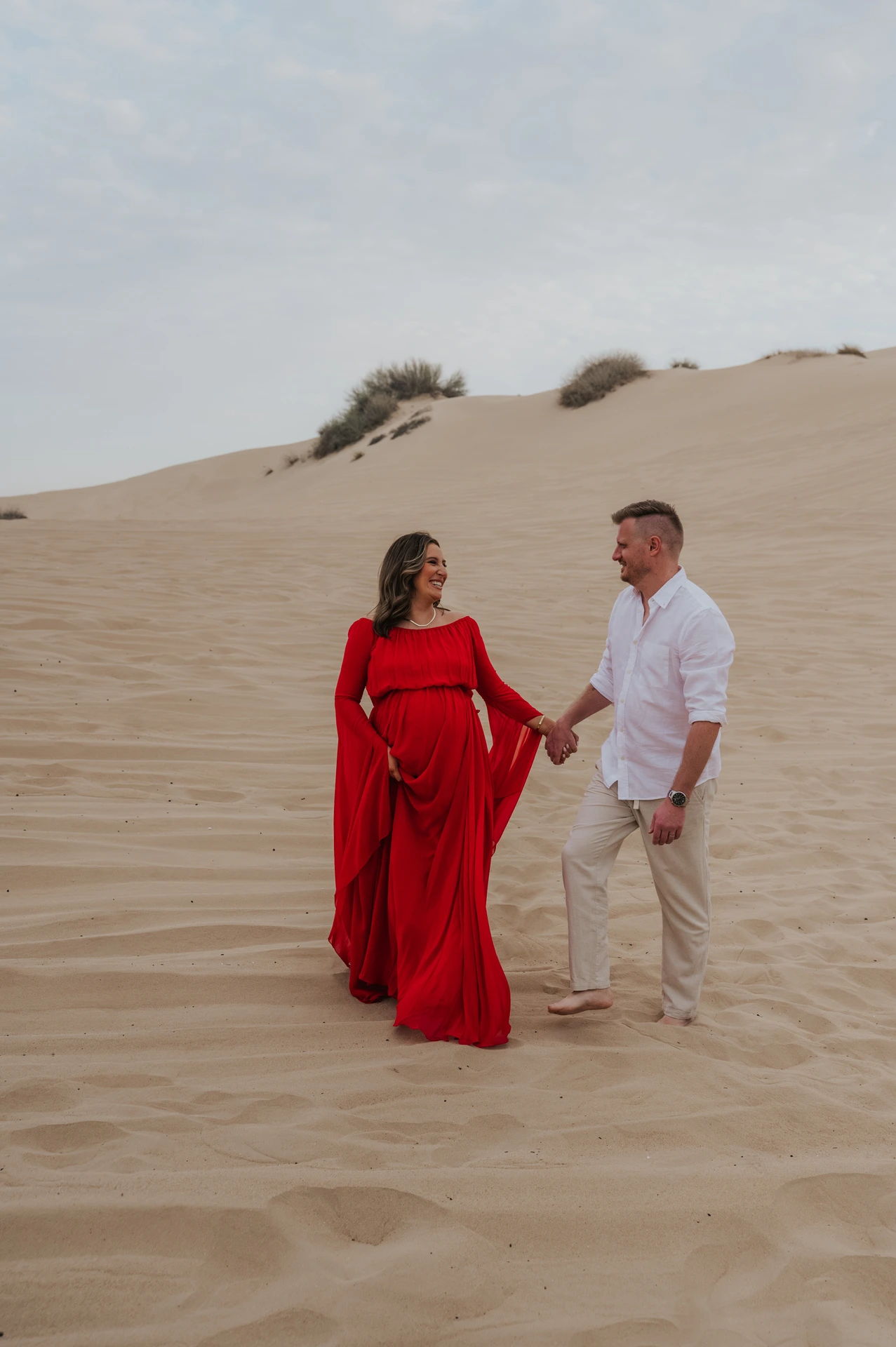 Pregnant woman in flowing red dress and partner hold hands on Dubai desert dunes during a maternity photoshoot.