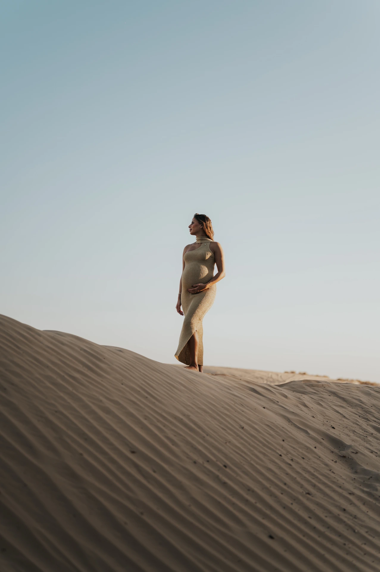 Pregnant woman in a fitted dress stands on sunlit Dubai desert dunes during a serene maternity photoshoot at golden hour.