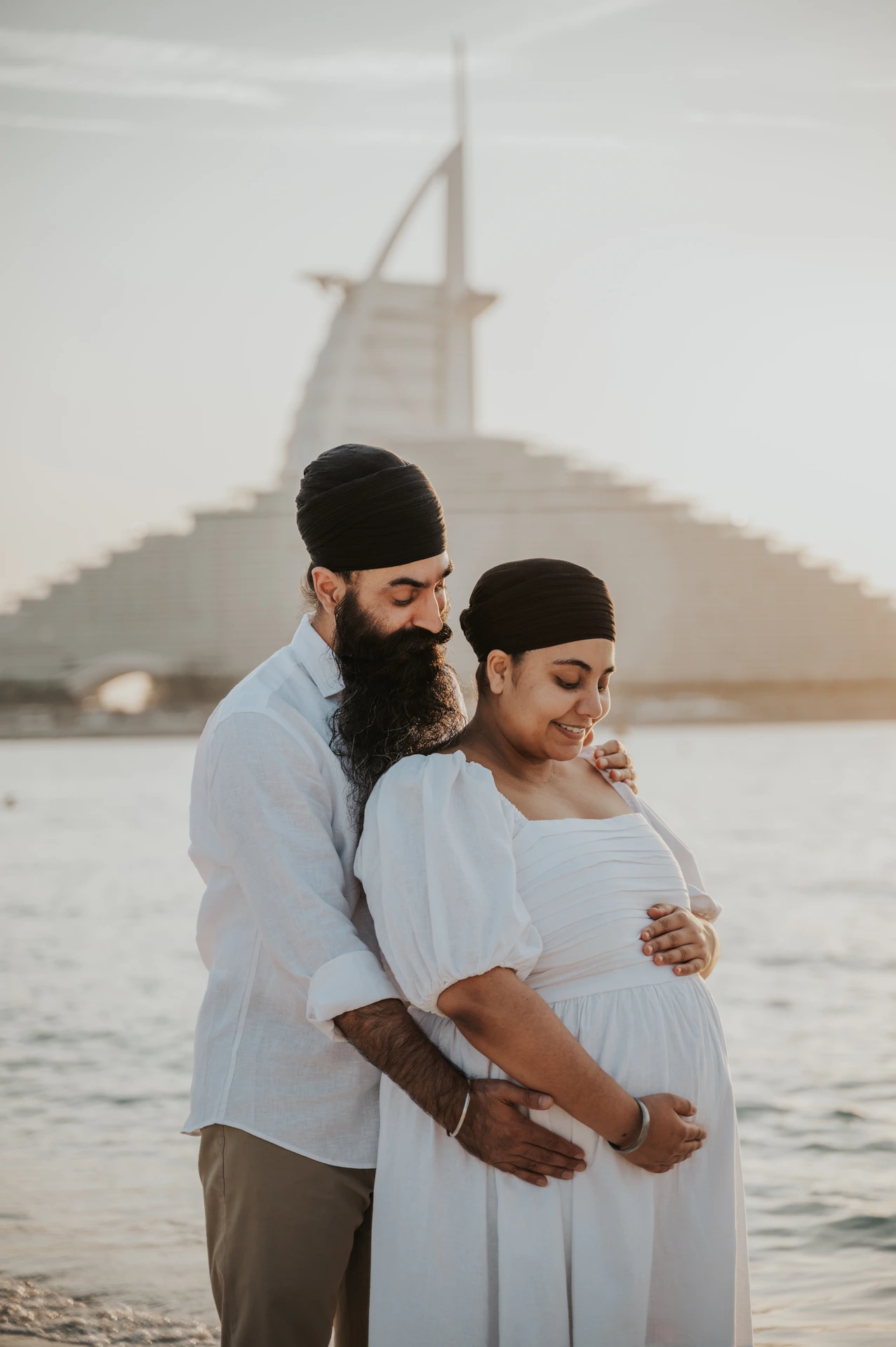 An expecting couple in light outfits walks on a quiet Dubai beach in the morning for a calm babymoon photoshoot.