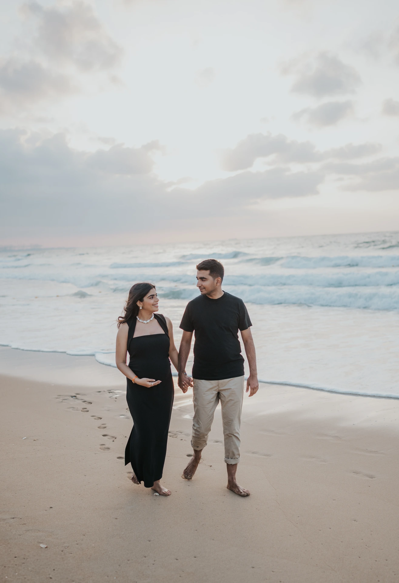 An expecting couple in dark outfits walks together on the Dubai beach at sunset during a babymoon session.