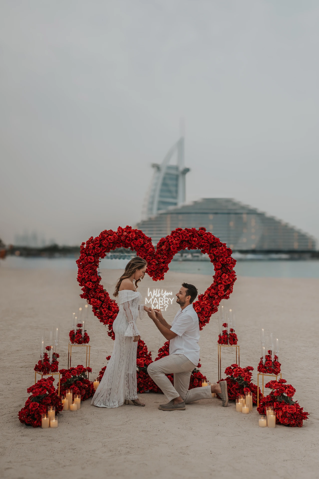 A proposal setup with a red floral heart arch and candles on the beach, with Burj Al Arab in the background.