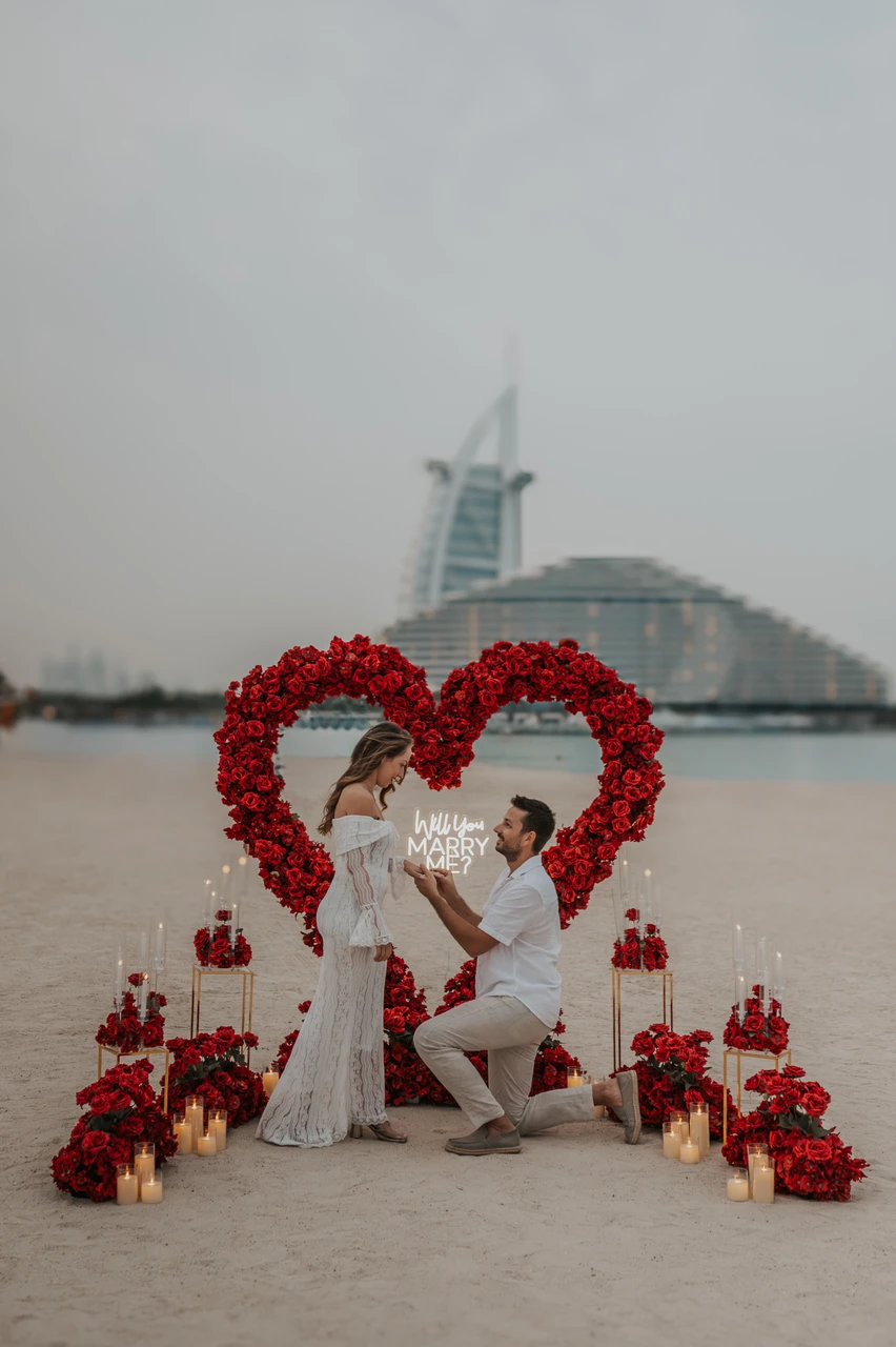 Beach proposal Dubai - Floral heart setup at Burj Al Arab A proposal setup with a red floral heart arch and candles on the beach, with Burj Al Arab in the background.