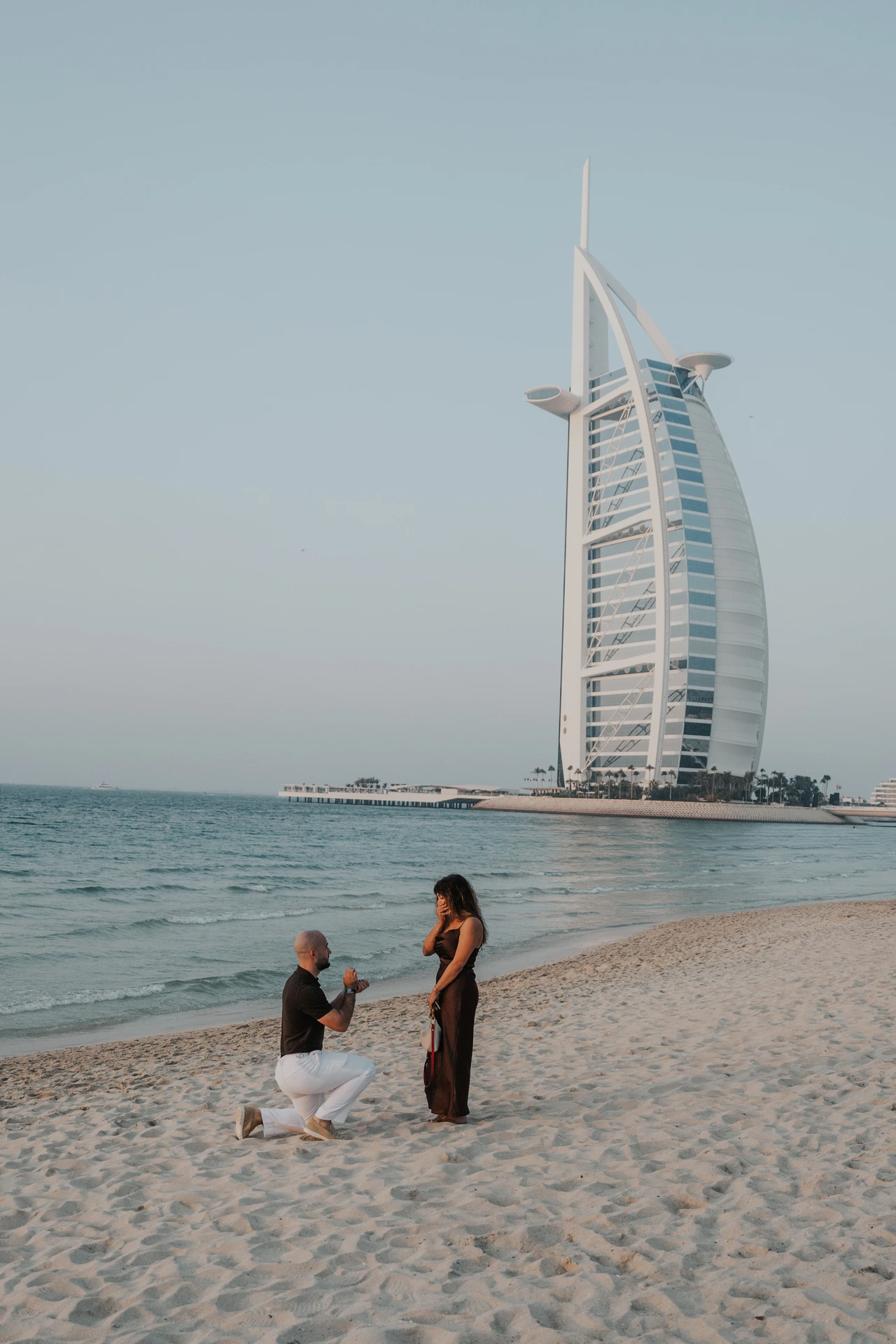 A man kneels to propose on a Dubai beach while his partner reacts, with Burj Al Arab towering in the background.