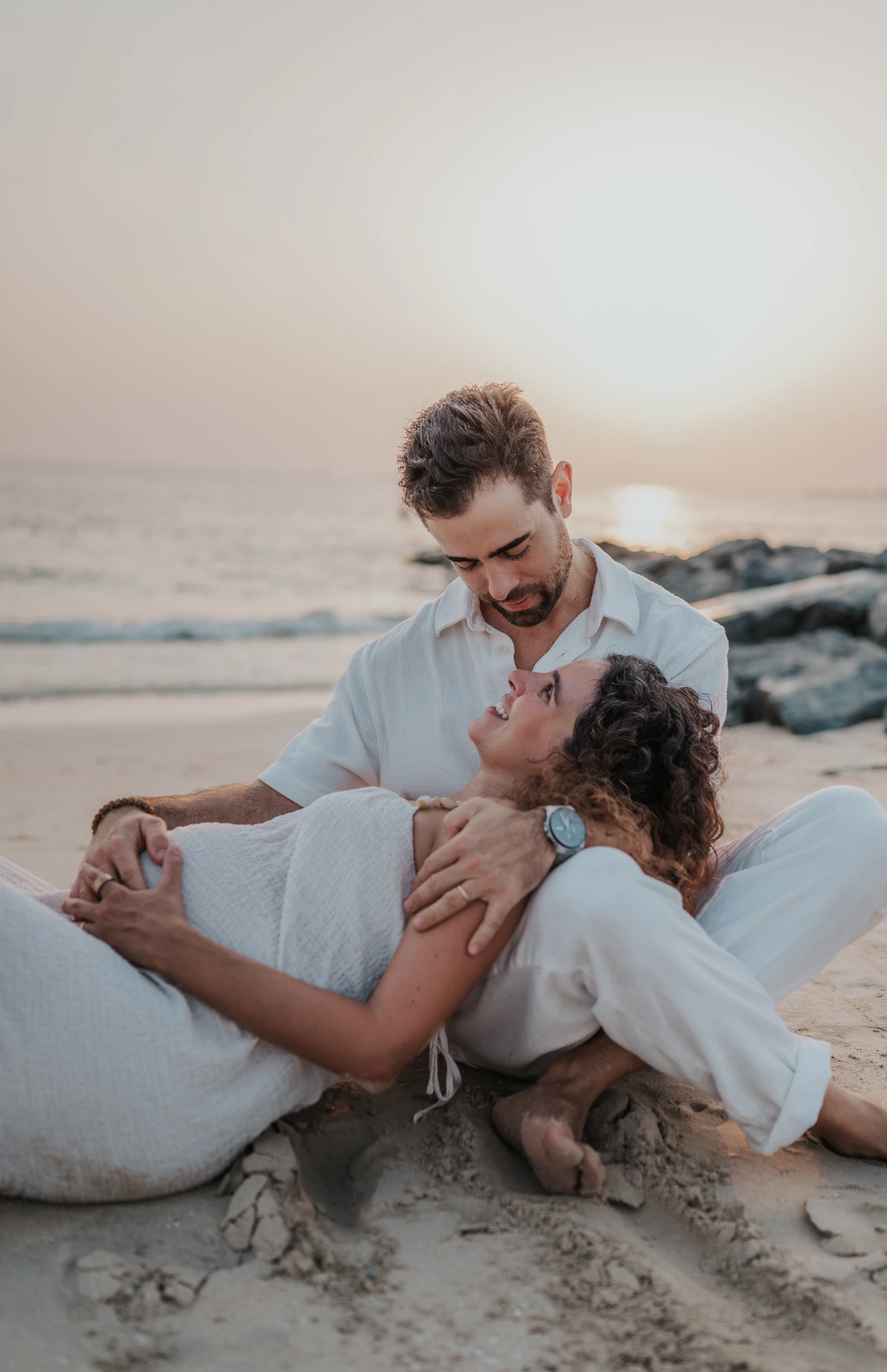 A couple cuddles on a Dubai beach while seated on the sand during a relaxed maternity session.