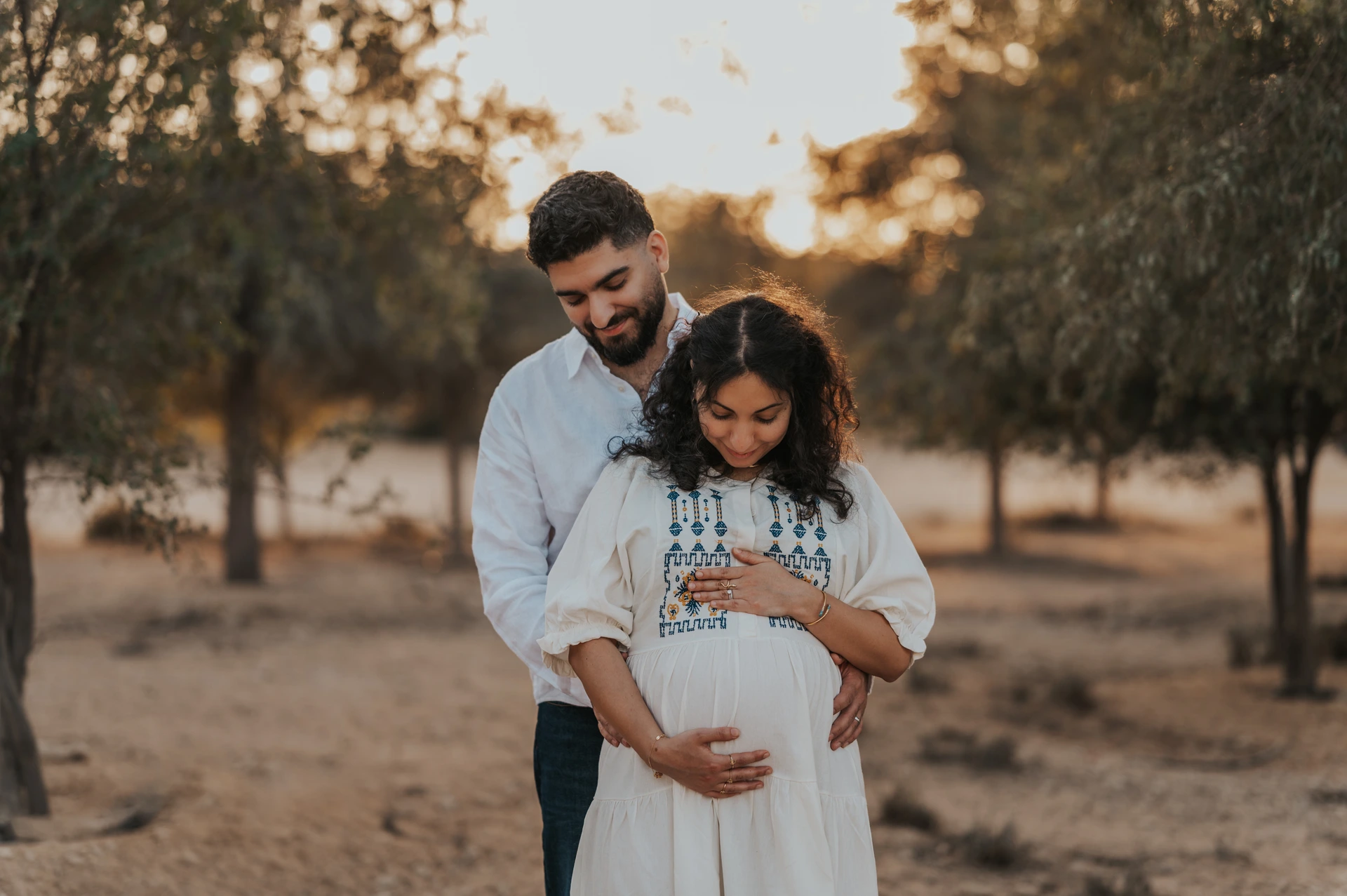 An expecting couple embraces in warm evening light in a park-like outdoor location in Dubai.