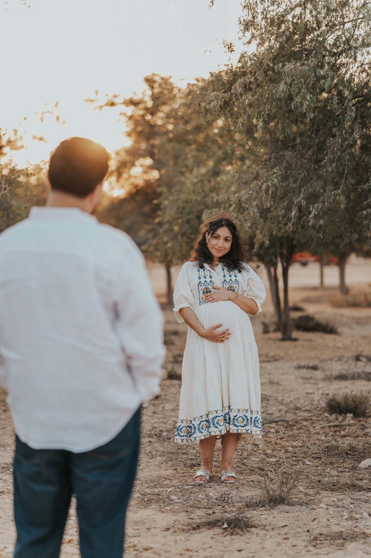 A pregnant woman in a white dress stands outdoors at sunset while her partner approaches from behind.