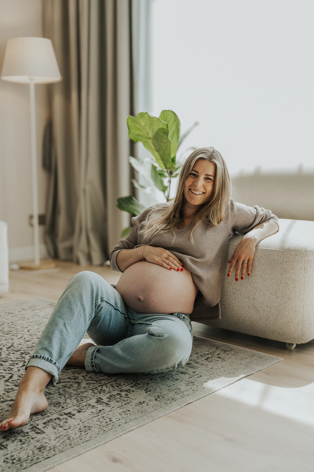 A pregnant woman relaxes on a sofa at home in a soft natural-light indoor maternity portrait.