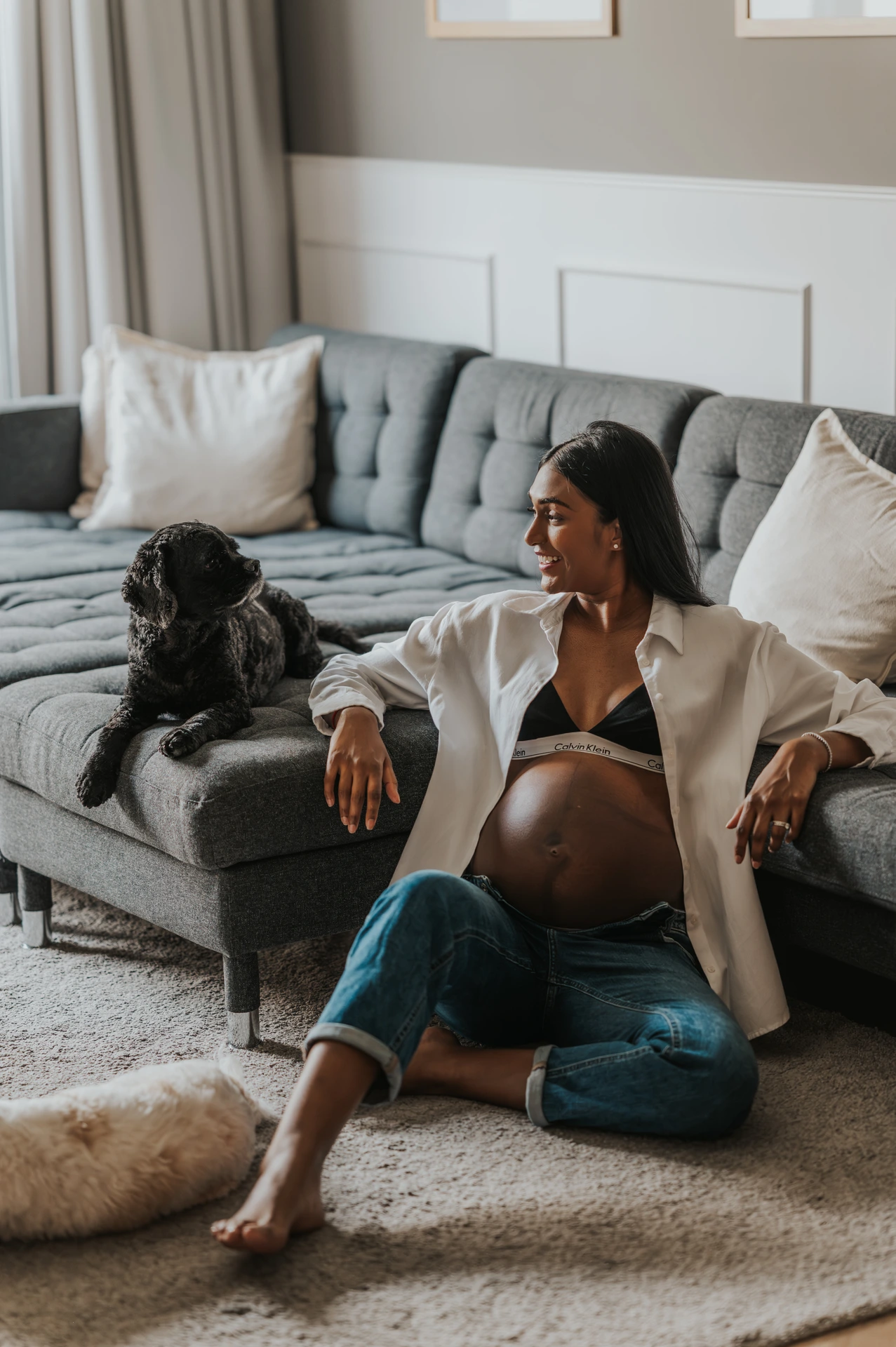 A pregnant woman sits on a couch with her dog during a relaxed lifestyle maternity photoshoot at home.