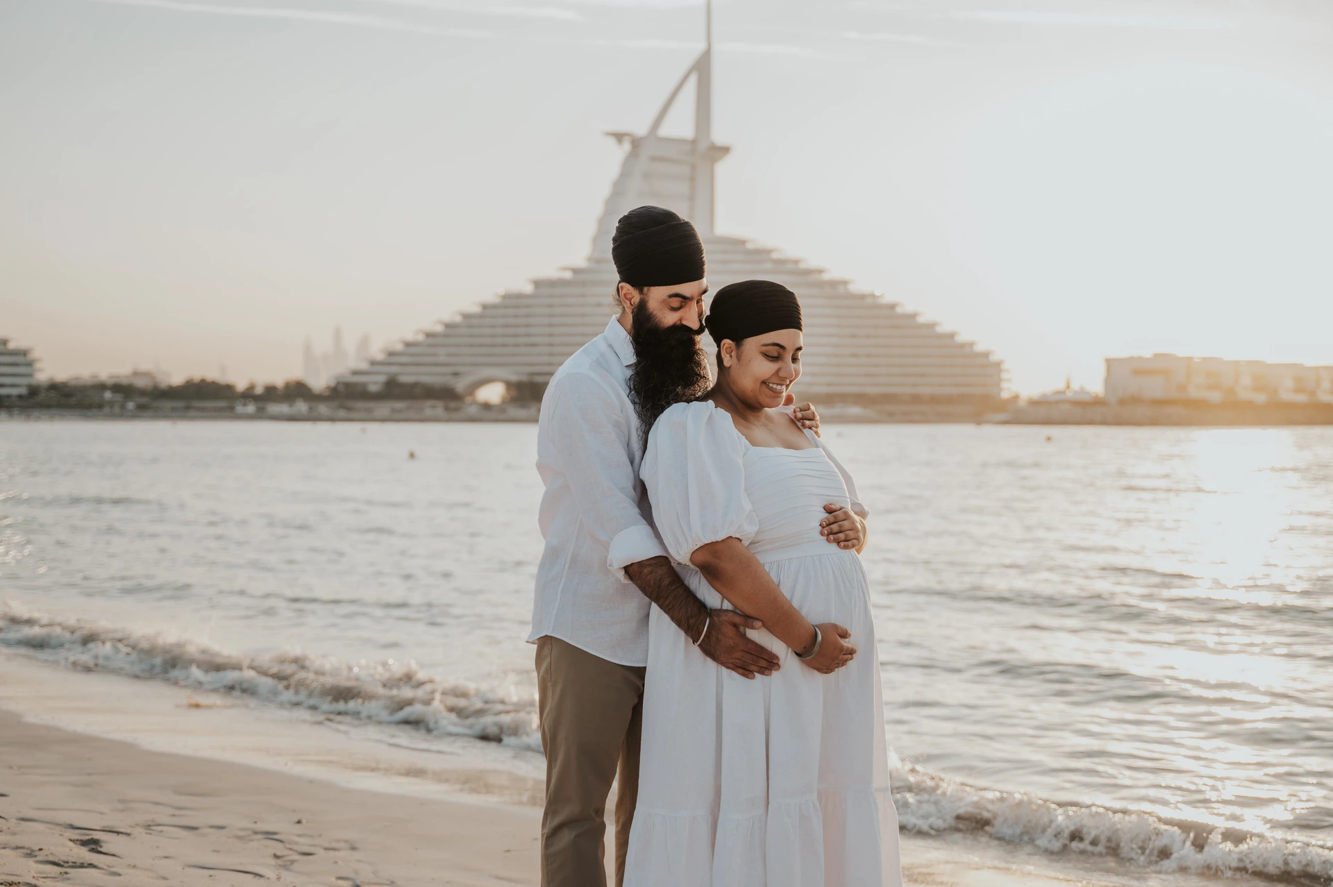 A partner is embracing an expecting mother on Dubai beach with Burj Al Arab in view during a maternity photoshoot portrait.