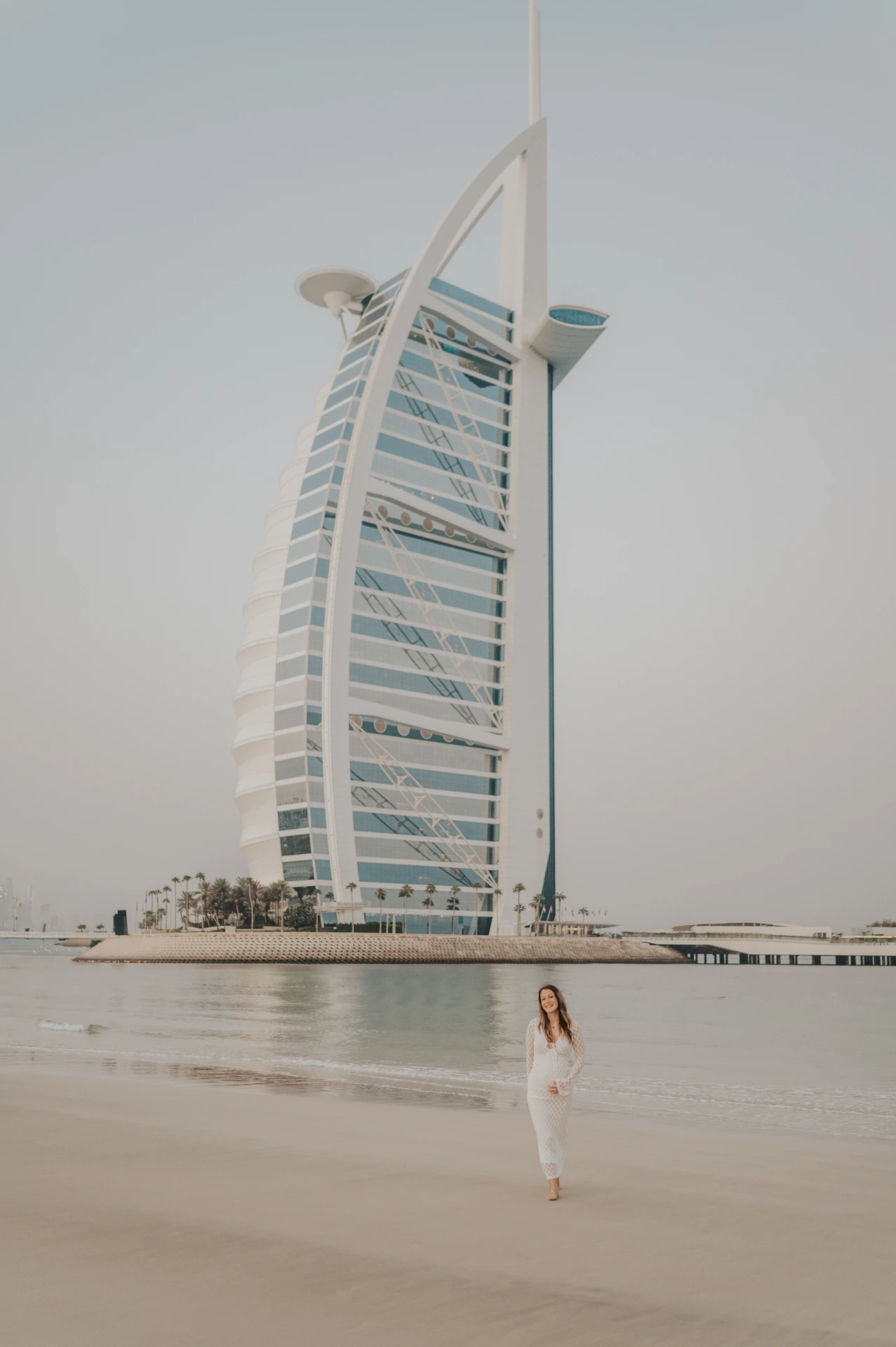A maternity couple stands near the waterline in Dubai with Burj Al Arab in frame during a relaxed beach photoshoot.