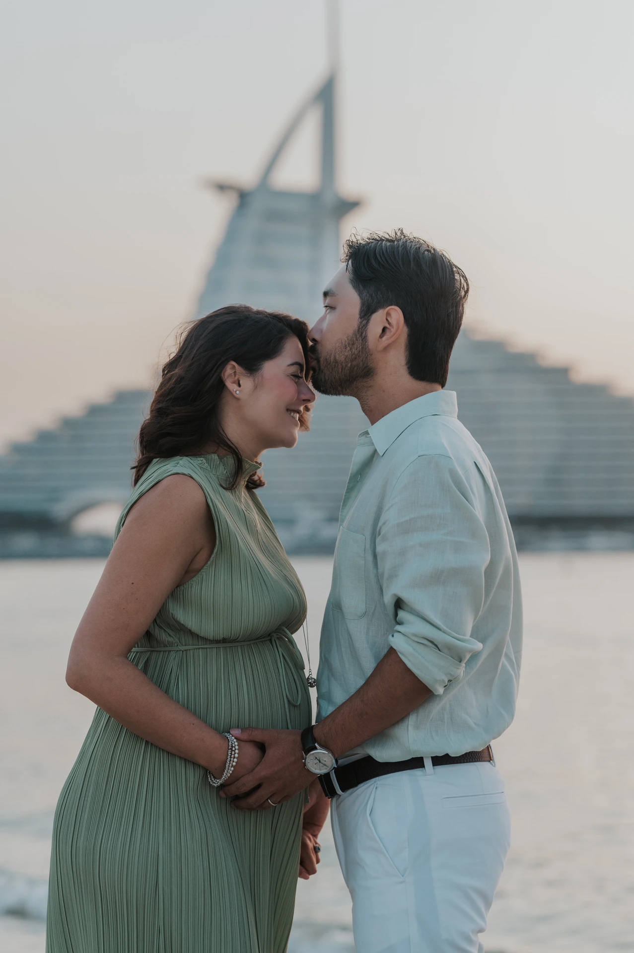 An expecting couple touches foreheads on Dubai beach with Burj Al Arab behind them during an intimate maternity photoshoot moment.
