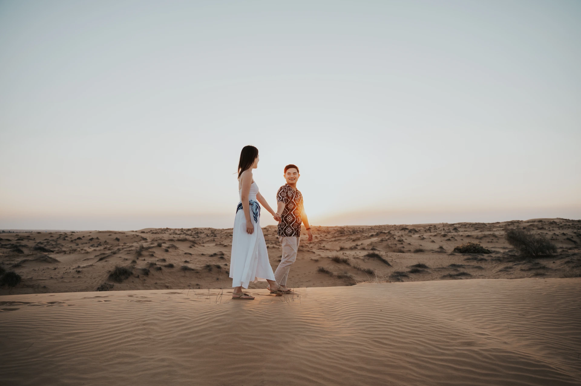 A couple walks through Dubai dunes in an engagement photoshoot, with natural movement and neutral styling suited to open desert compositions.