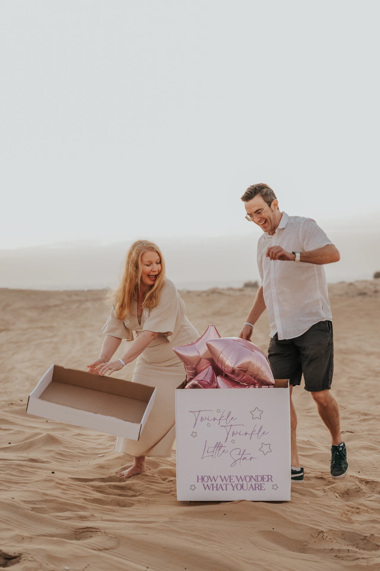 A couple opens a surprise box with pink star balloons on Dubai desert sand during a candid gender reveal photoshoot.