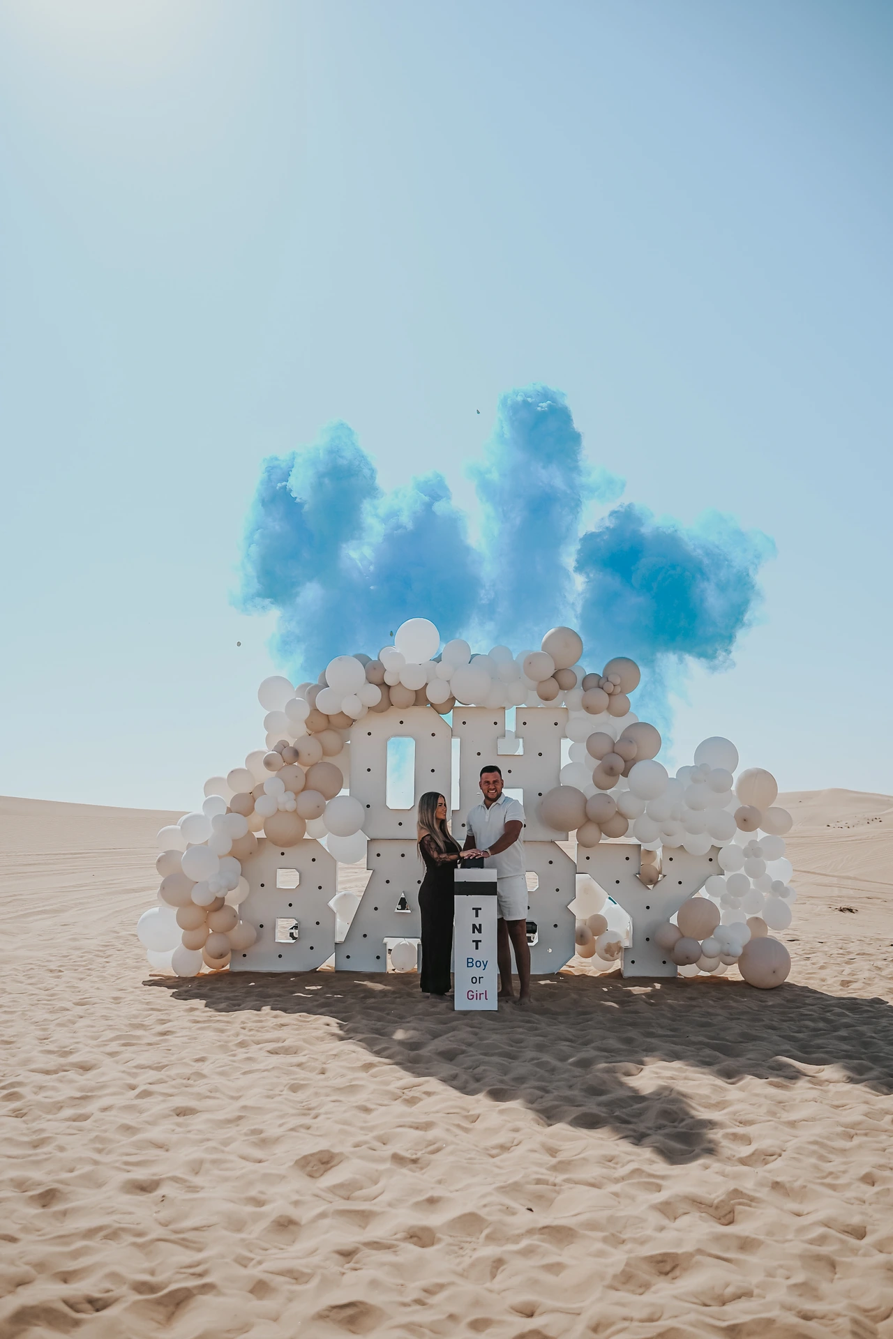 A couple stands in front of BABY letters and a balloon arch in Dubai desert as blue smoke rises during a gender reveal photoshoot.