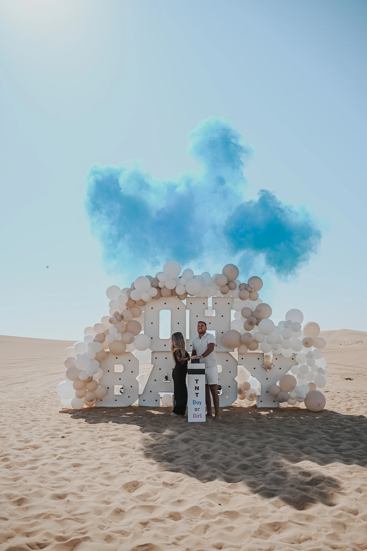 A couple poses at a BABY letter setup in Dubai desert with blue smoke and balloons during a dramatic gender reveal photoshoot.