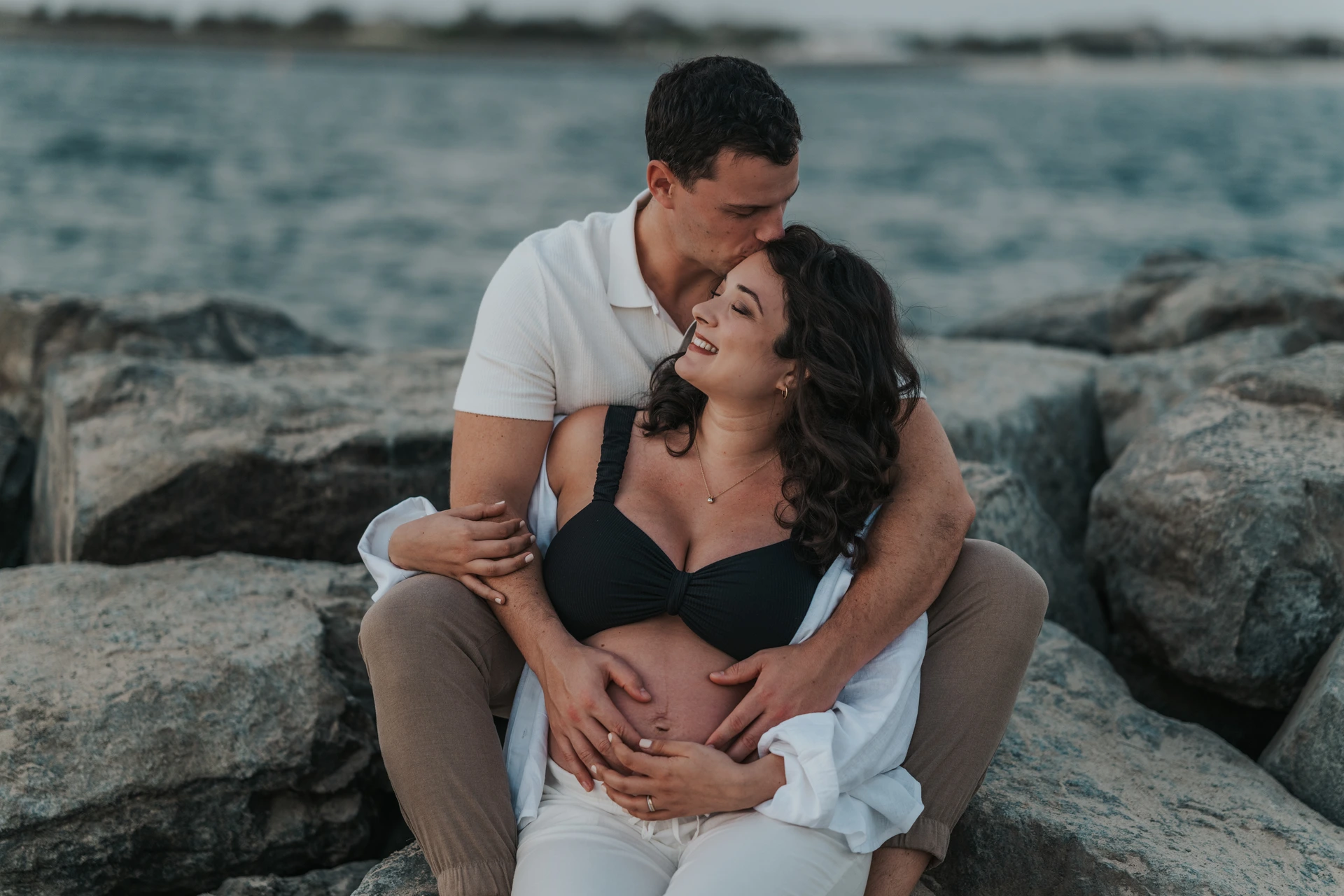 A couple sits together on shoreline rocks in Dubai during a maternity shoot, wearing fitted neutrals and open layers suited for textured coastal backgrounds.