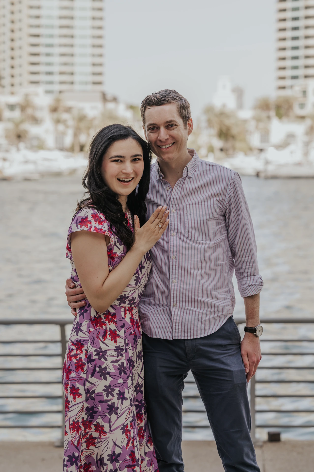 A smiling couple embraces on Dubai Marina promenade after a proposal, with waterfront towers softly blurred behind.