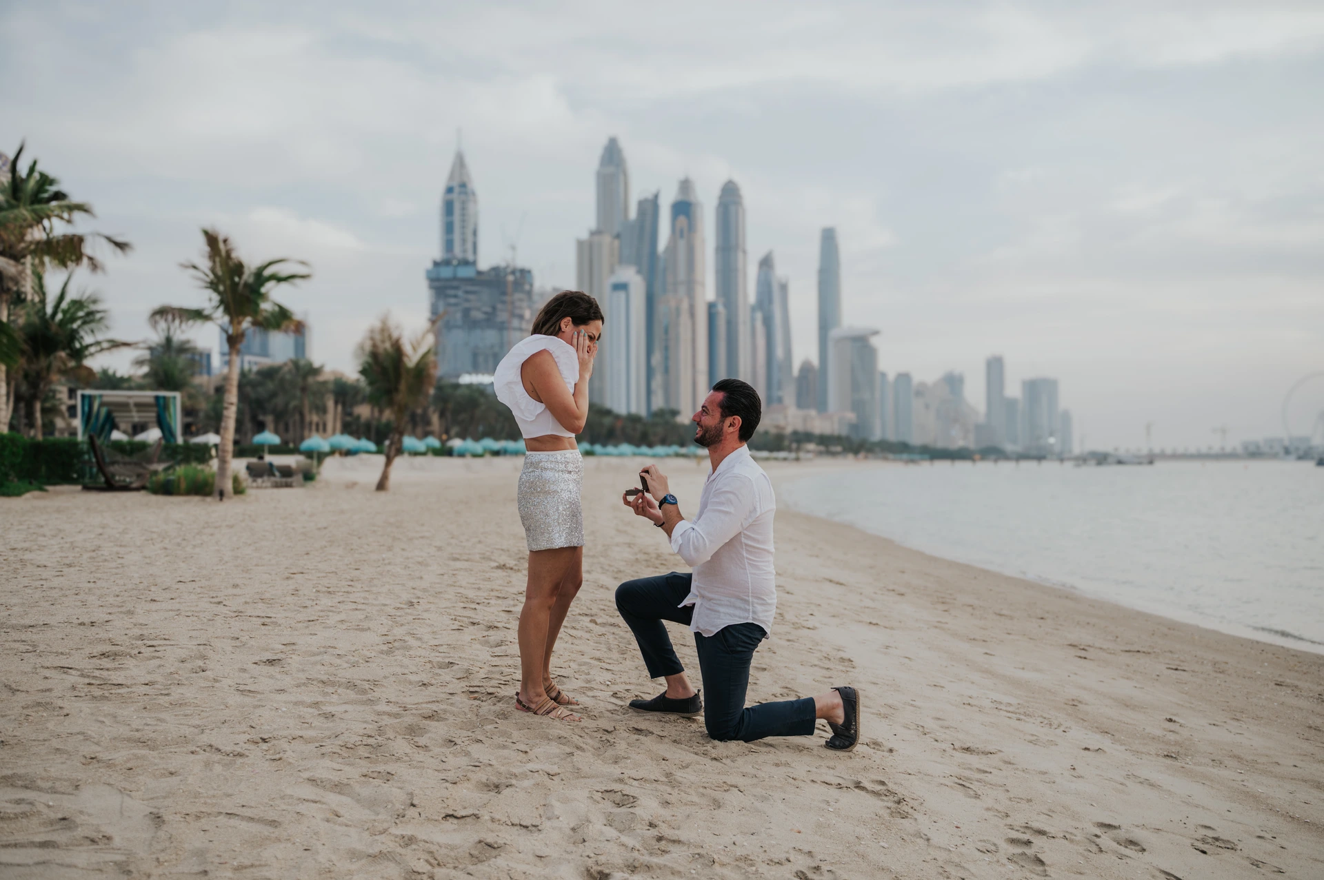 A man kneels on the beach and proposes as Dubai Marina skyline rises behind the couple at sunset.