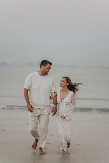 Maternity photoshoot beach vs desert - white outfit beach couple An expecting couple in white outfits stands by the sea in Dubai during a maternity photoshoot, creating a clean and timeless beach portrait.