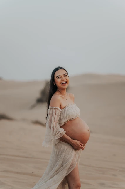 Maternity photoshoot beach vs desert - desert solo maternity smile A pregnant woman smiles on Dubai desert dunes during a maternity photoshoot, highlighting a solo portrait style with soft natural desert tones.