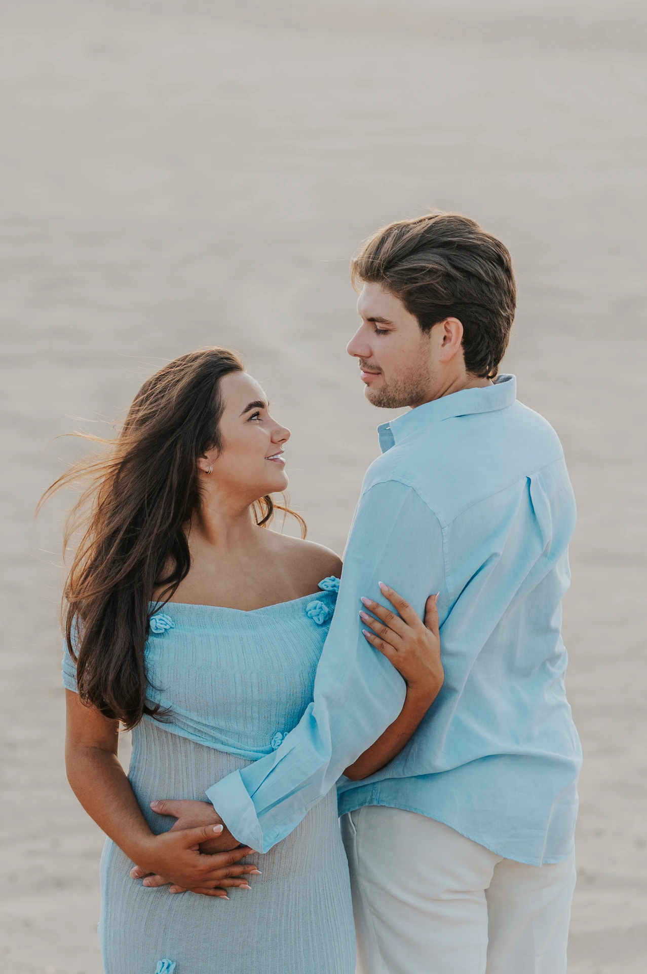An expecting couple in blue and white outfits shares a close look on Dubai dunes during a maternity photoshoot at golden hour.