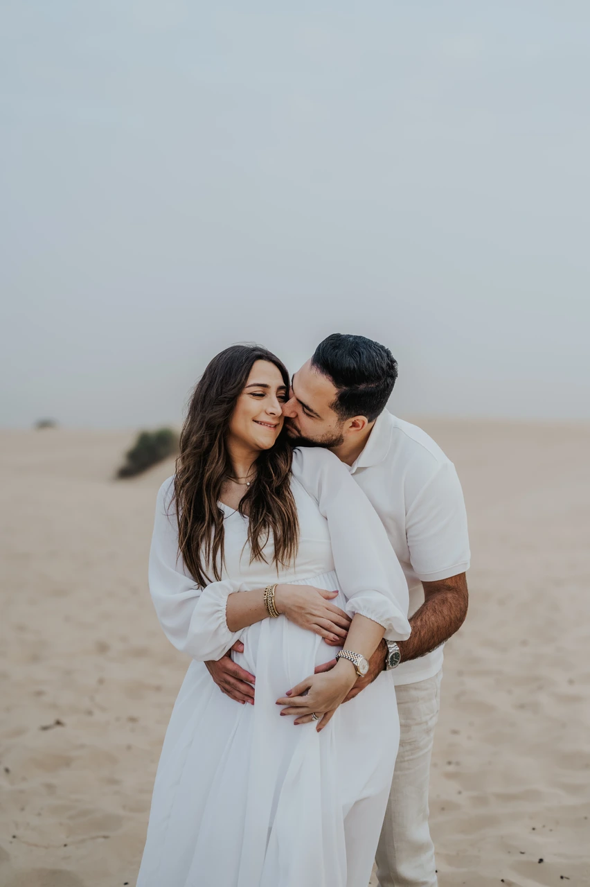 Planning desert maternity photoshoot Dubai - kiss on cheek A partner kisses an expecting mother on the cheek on Dubai desert dunes during a maternity photoshoot with natural affectionate expression.