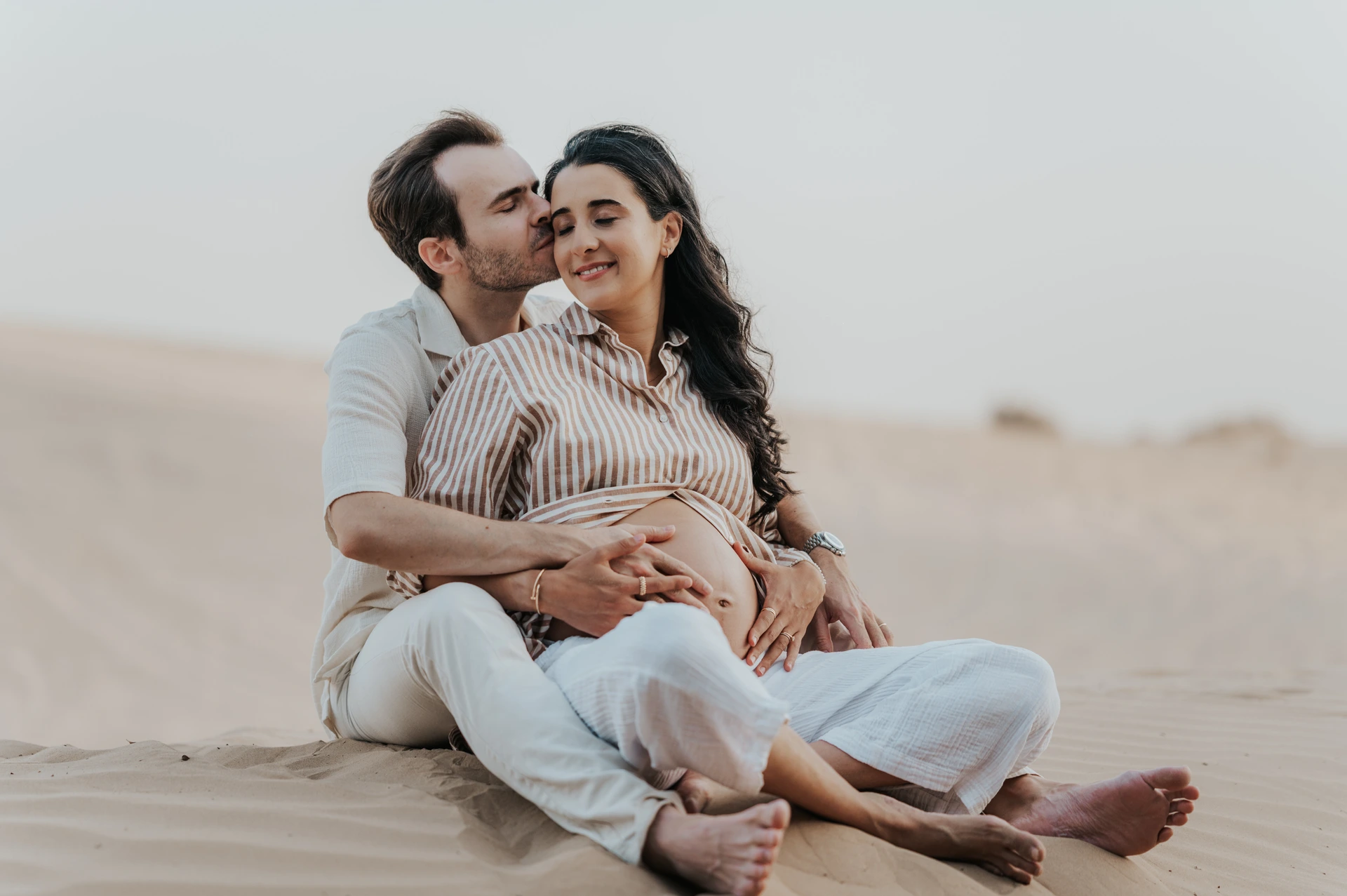An expecting couple sits together on Dubai sand during a maternity photoshoot, hugging closely in a calm storytelling composition.