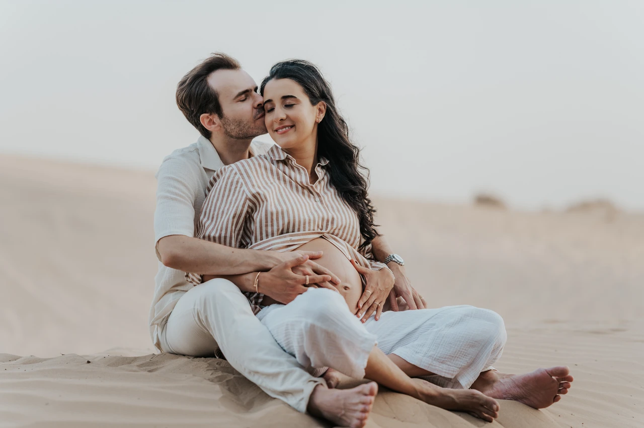 Planning desert maternity photoshoot Dubai - seated embrace An expecting couple sits together on Dubai sand during a maternity photoshoot, hugging closely in a calm storytelling composition.