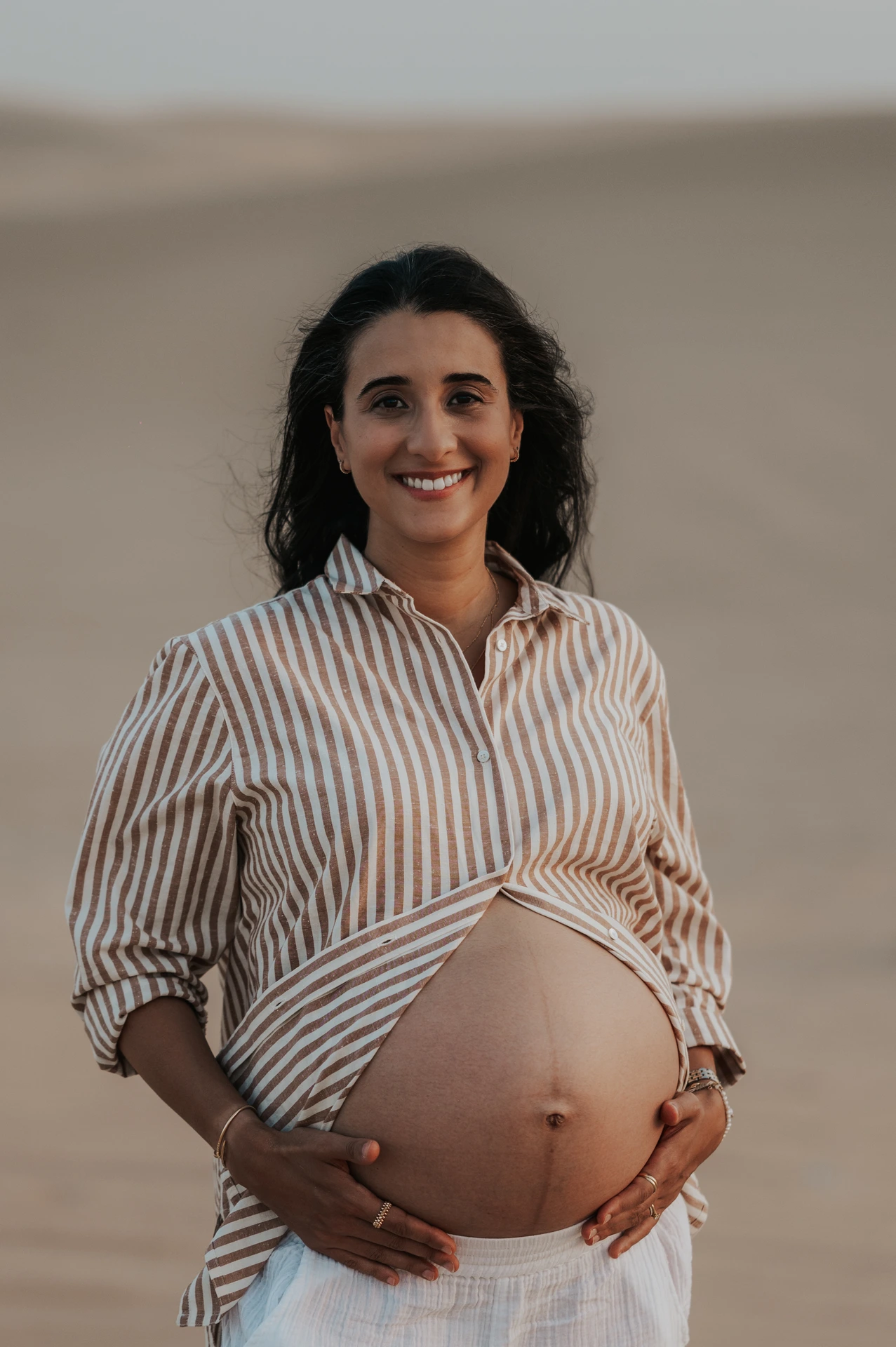 An expecting mother smiles while holding her striped shirt at Dubai dunes, revealing her bump during a candid maternity photoshoot.
