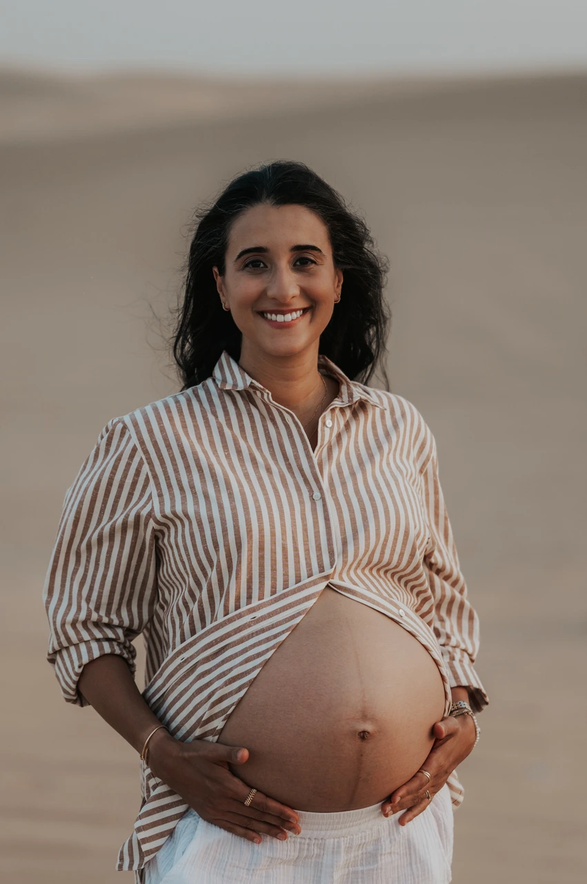 Planning desert maternity photoshoot Dubai - striped shirt detail An expecting mother smiles while holding her striped shirt at Dubai dunes, revealing her bump during a candid maternity photoshoot.