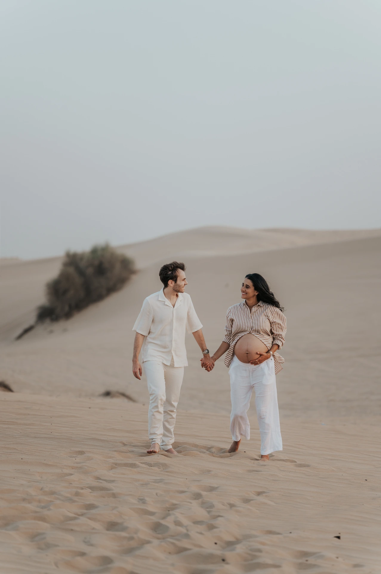 An expecting couple walks hand in hand on Dubai desert dunes during a maternity photoshoot, smiling as they move through soft evening light.