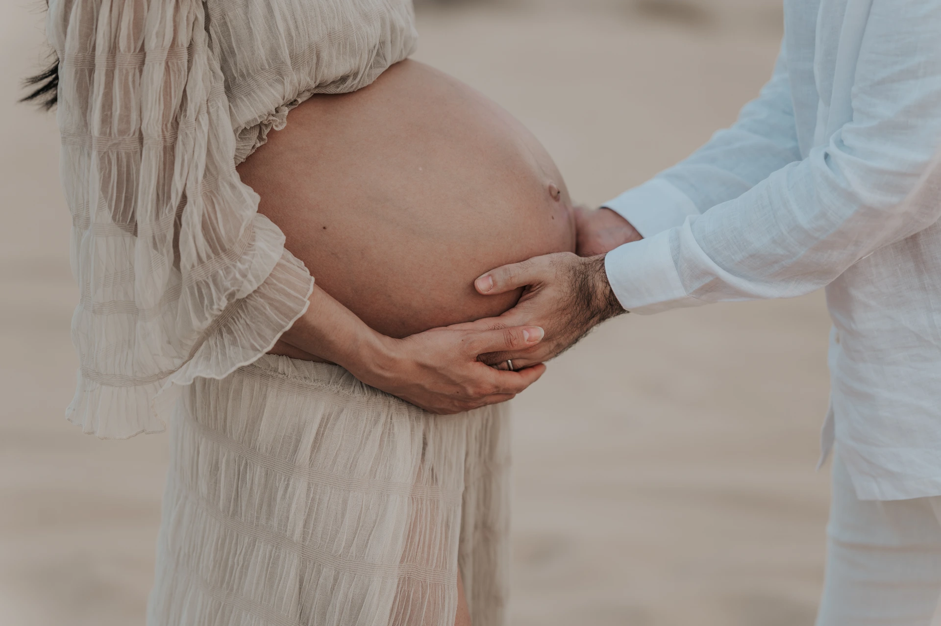 Close-up of hands around a pregnant belly on a Dubai beach during a maternity photoshoot, capturing intimate detail in a natural outdoor setting.