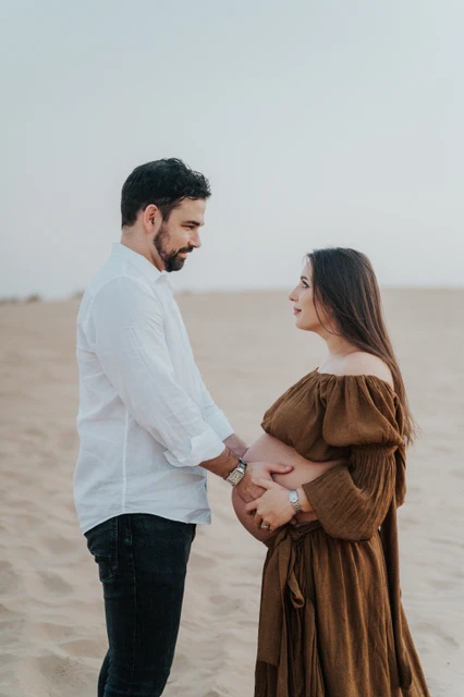 Planning desert maternity photoshoot Dubai - facing handhold An expecting couple faces each other and holds hands on Dubai desert dunes during a maternity photoshoot with warm natural light.