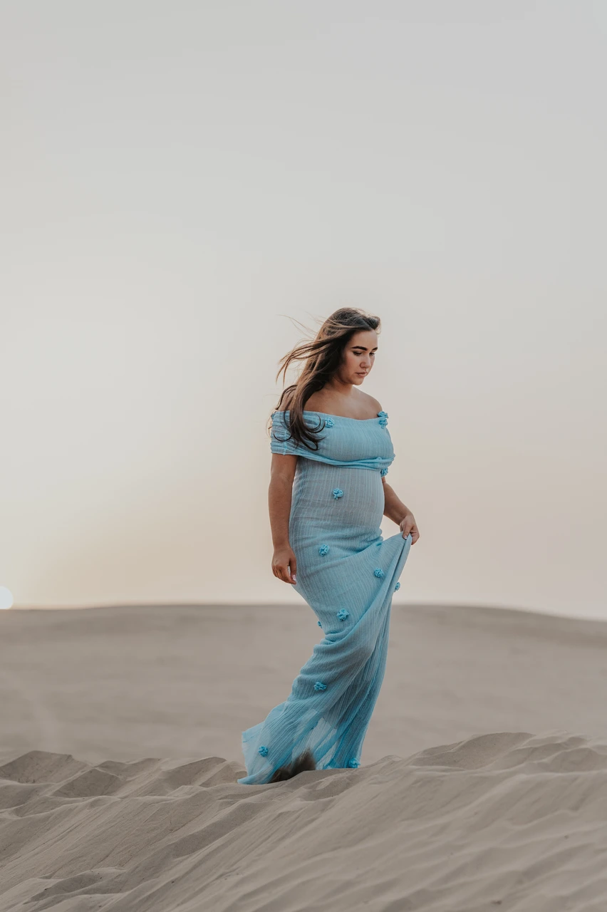 Planning desert maternity photoshoot Dubai - blue dress walk A pregnant woman in a blue dress walks across Dubai desert sand during a maternity photoshoot, creating gentle movement in fabric and posture.