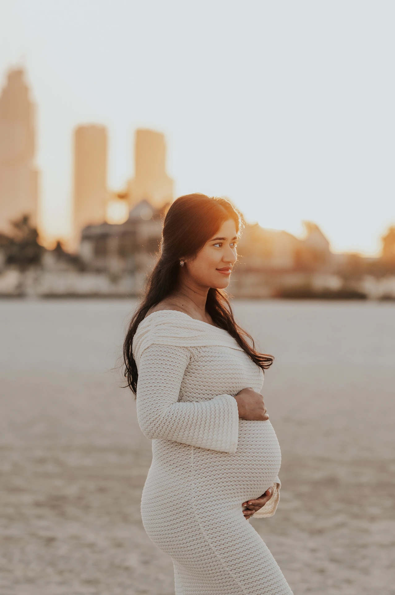 An expecting mother stands in profile on Dubai beach at golden hour during a maternity photoshoot, with soft city skyline blur behind her.