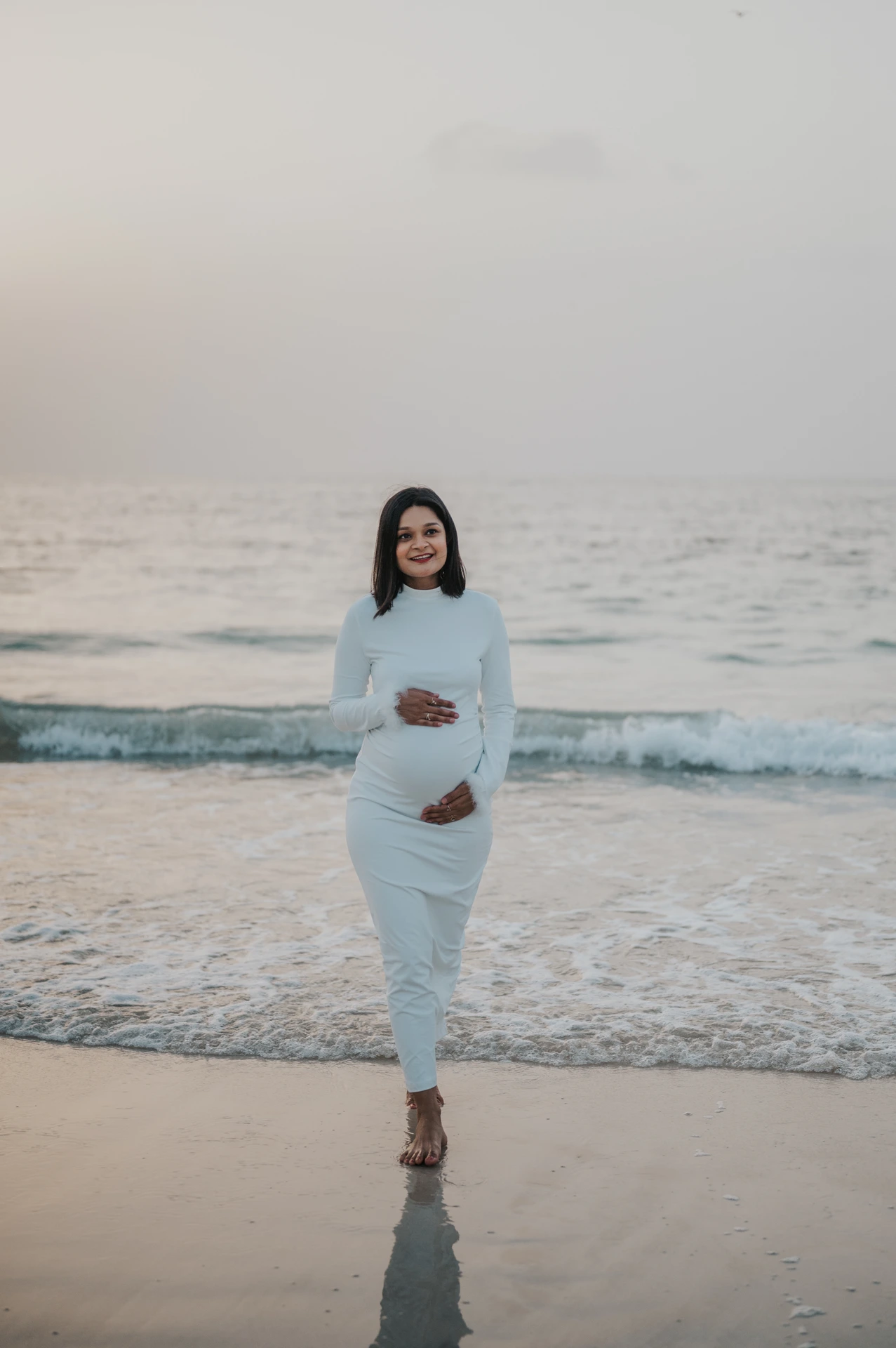 A pregnant woman poses in a white dress near the waterline in Dubai during a maternity photoshoot, with gentle waves rolling behind her.
