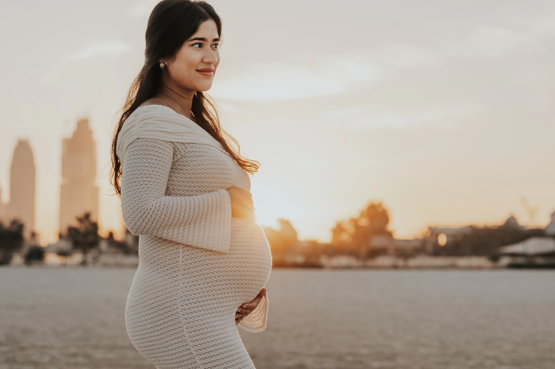 A pregnant woman gently holds her baby bump on Dubai beach at sunset during a maternity photoshoot with warm light and a calm backdrop.
