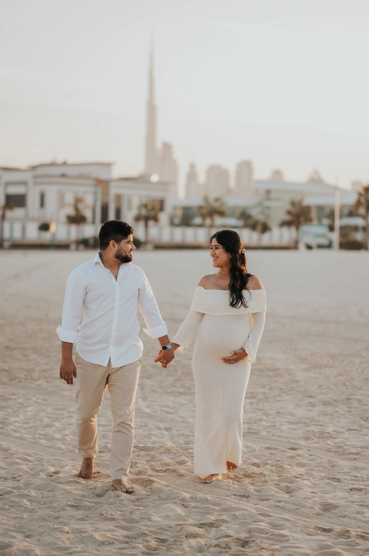 An expecting couple walks hand in hand across Dubai beach during a maternity photoshoot, smiling as evening light softens the scene.