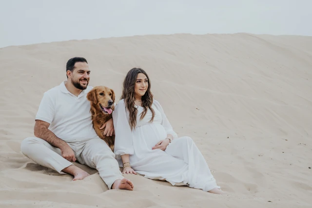 An expecting couple sits together on Dubai desert dunes with their dog during a maternity photoshoot with a pet, creating a calm family portrait.