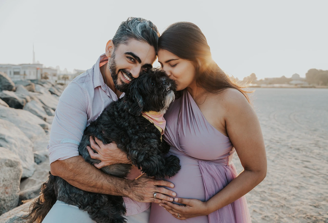 Maternity photoshoot with your pet - Dubai beach family cuddle A pregnant couple hugs their black dog on a Dubai shoreline during a maternity photoshoot with a pet, capturing a close and emotional family moment.