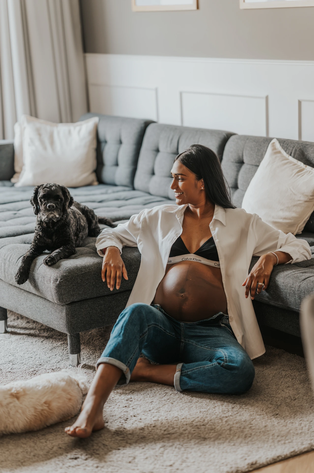 A pregnant woman sits on a sofa with her dog in Dubai during a maternity photoshoot with a pet, capturing a cozy and candid indoor frame.