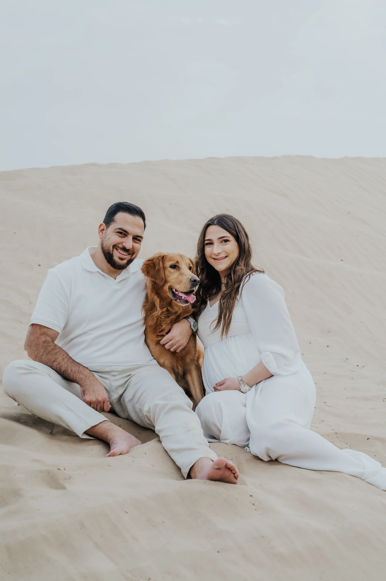 An expecting couple sits with their dog on Dubai dunes during a maternity photoshoot with a pet, creating a warm and balanced family portrait.