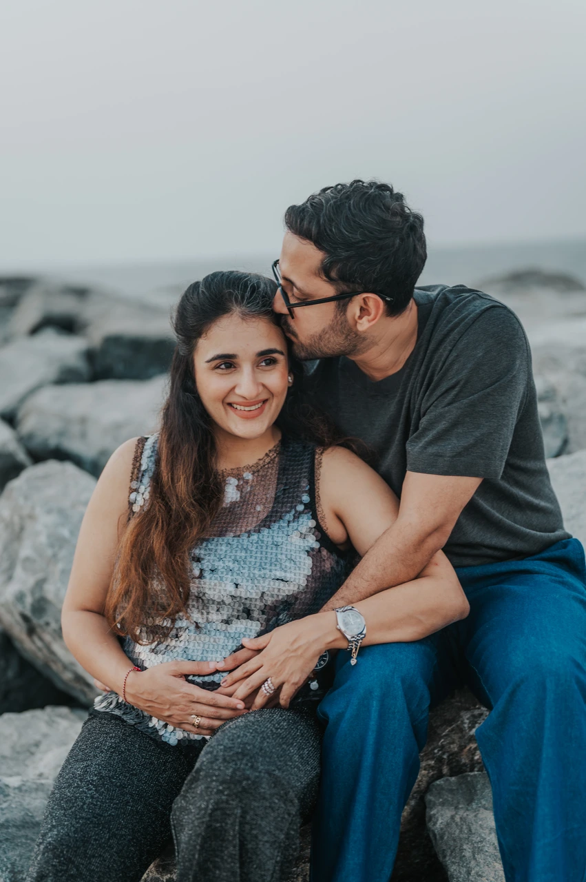 Maternity Photoshoot Dubai - couple on seaside rocks An expecting couple sits on seaside rocks in Dubai during a maternity photoshoot, smiling closely while the partner kisses her temple.
