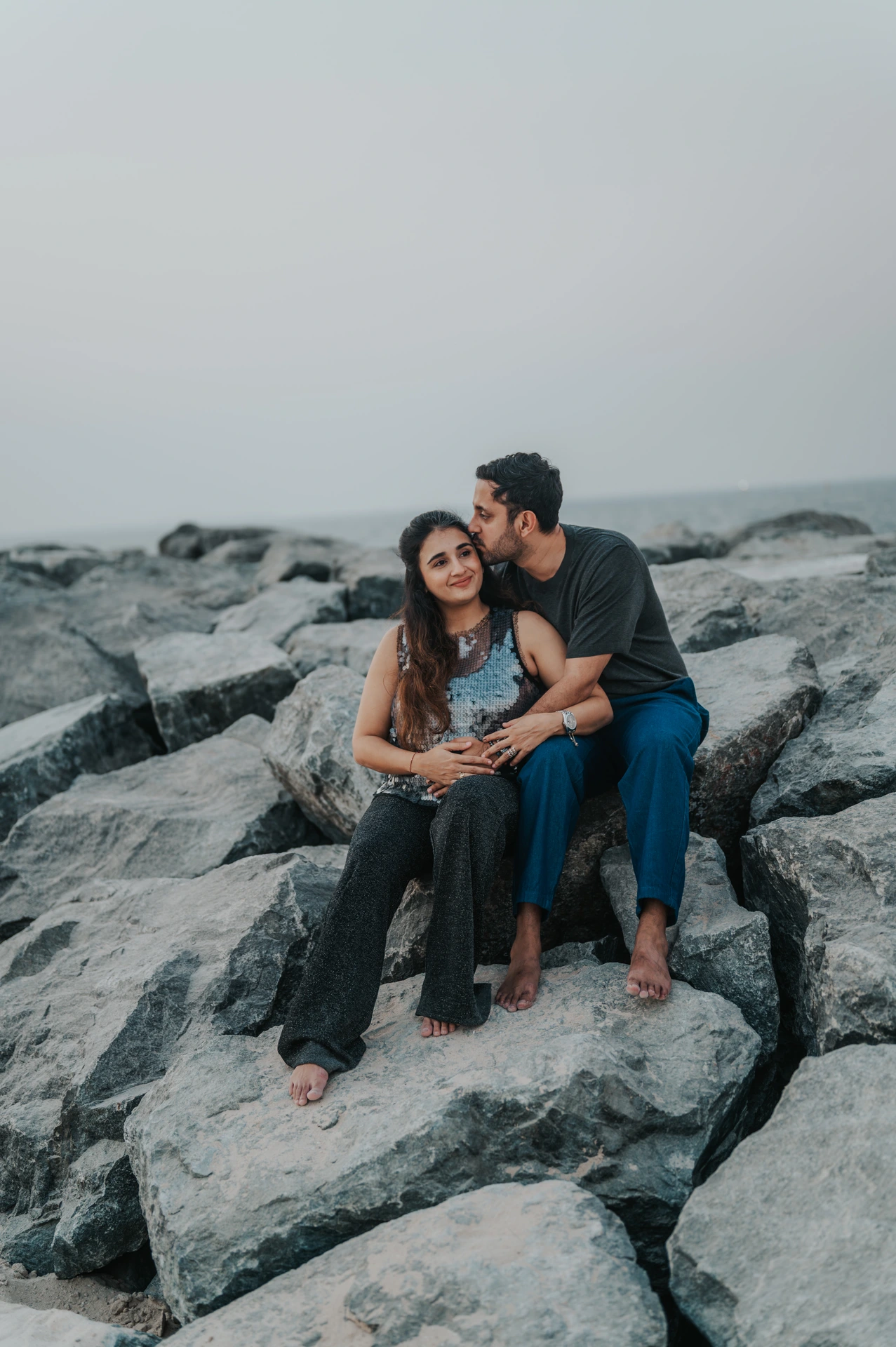 An expecting couple sits together on large coastal rocks in Dubai during a maternity photoshoot, creating a relaxed wide portrait by the sea.