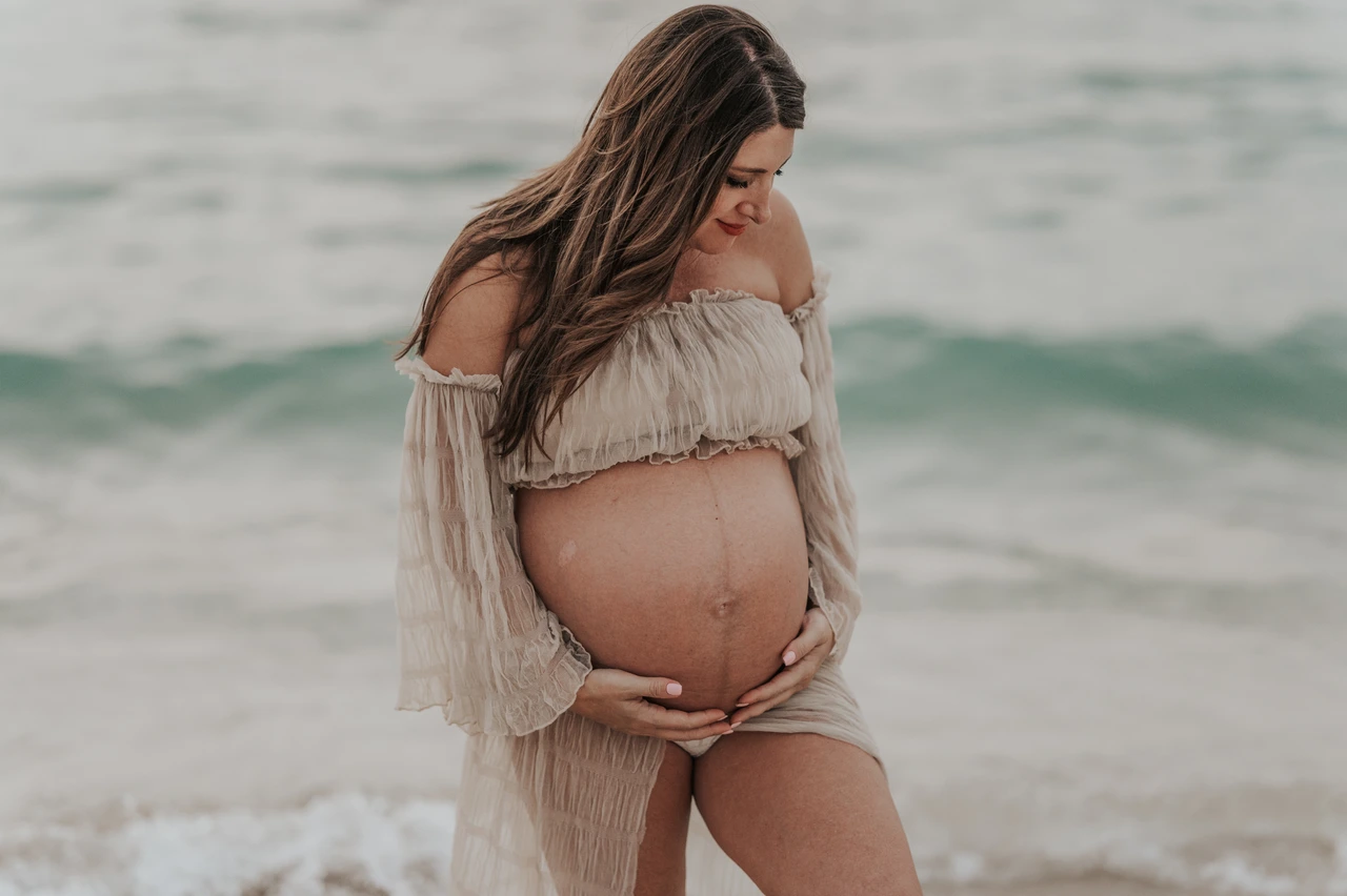 Maternity Photoshoot Dubai - shoreline bump portrait A pregnant woman cradles her baby bump at Dubai beach during a maternity photoshoot, with ocean waves softly blurred in the background.