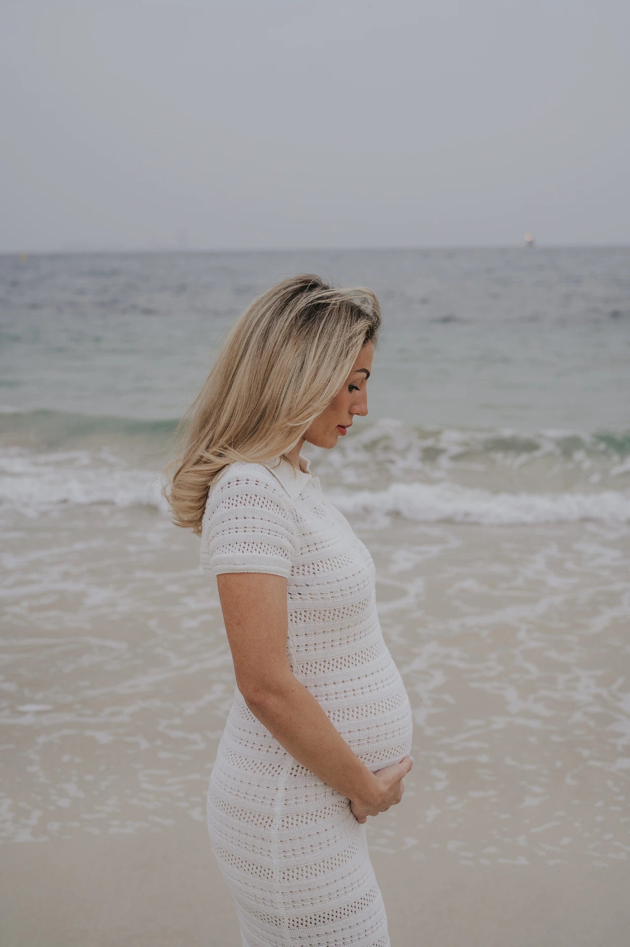 A pregnant woman stands in side profile at Dubai beach during a maternity photoshoot, holding her bump as waves break behind her.